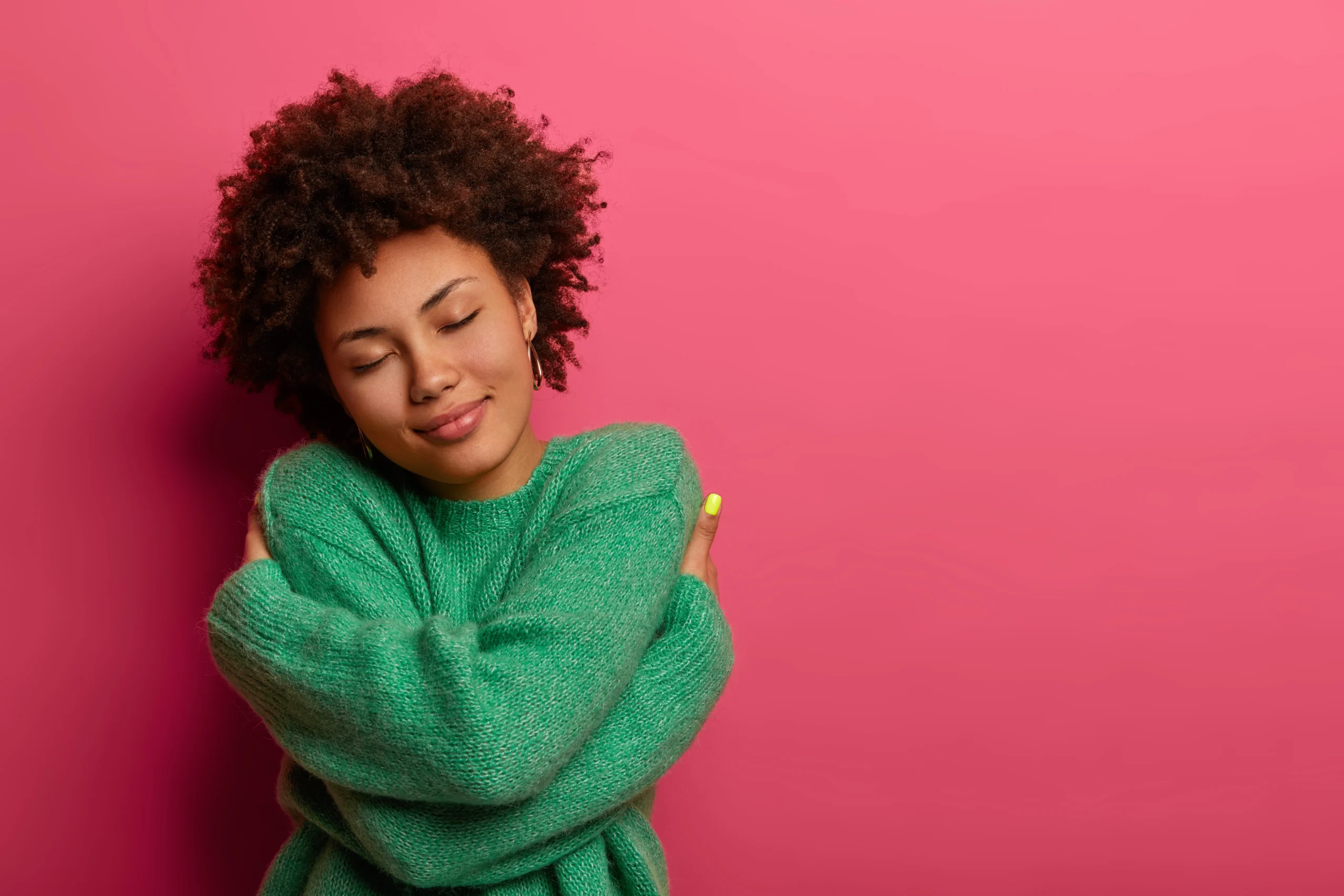 Woman hugging herself with eyes closed against pink background representing self-compassion and emotional wellness.