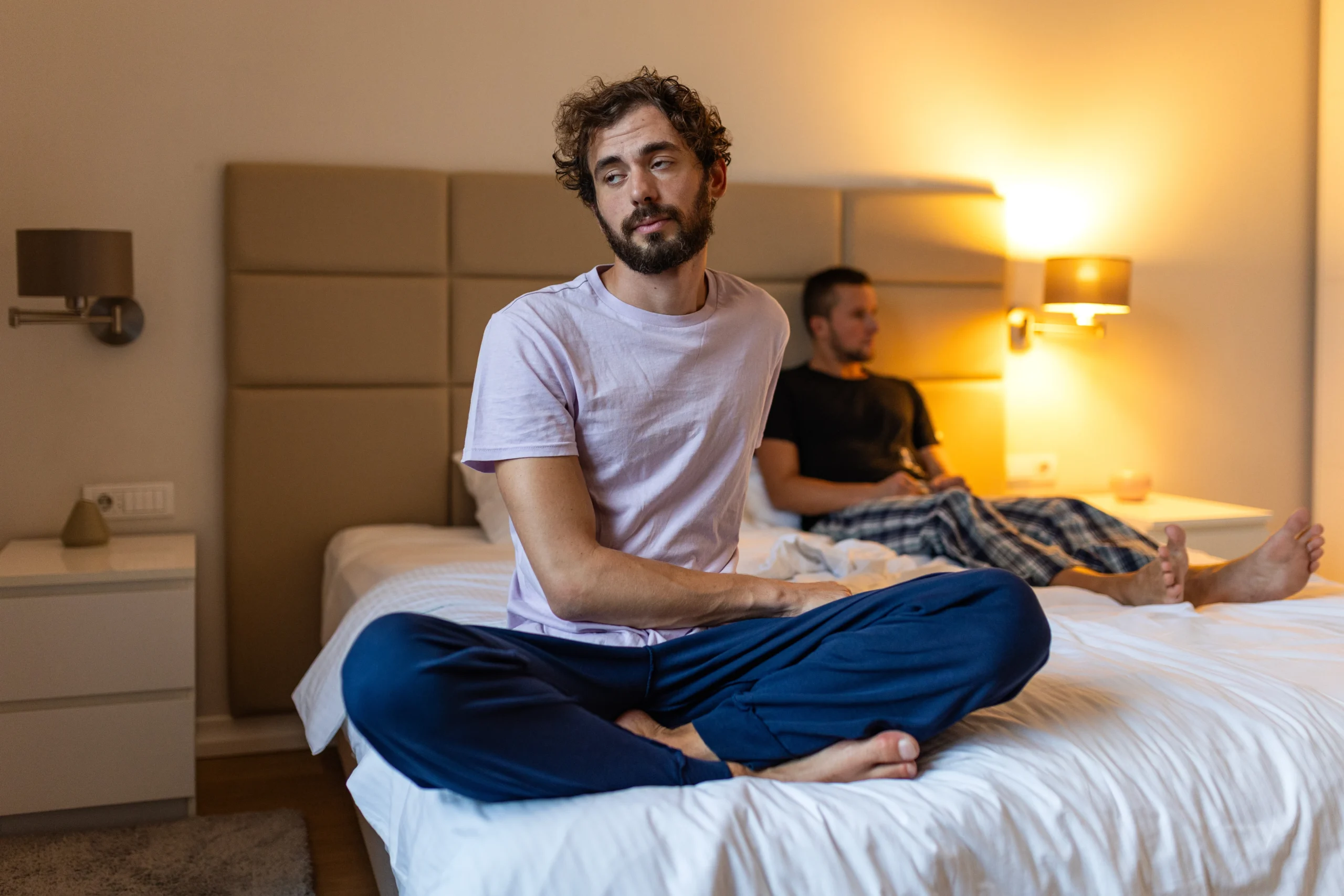 Two men sitting together in a bedroom, one appearing distressed and disconnected, representing the mental health challenges faced by LGBTQ individuals seeking affirming psychiatric care in Atlanta.