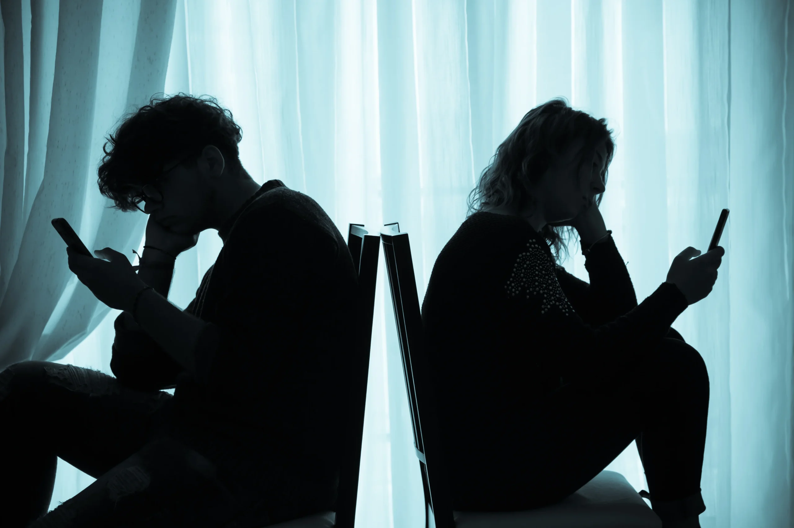 Two people sitting back to back in silhouette, each looking at a phone, representing the link between social media likes and depression discussed by Georgia Behavioral Health in Norcross, GA